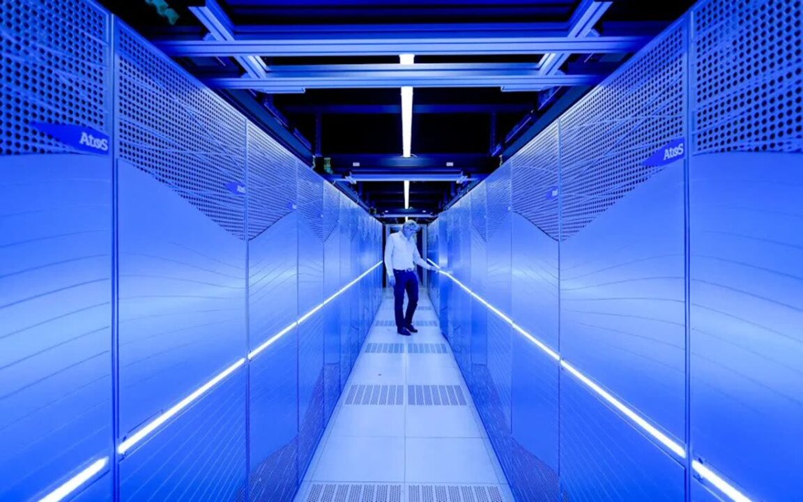 1000043378 Man in a white shirt walks down a narrow blue-lit data center aisle between server racks.