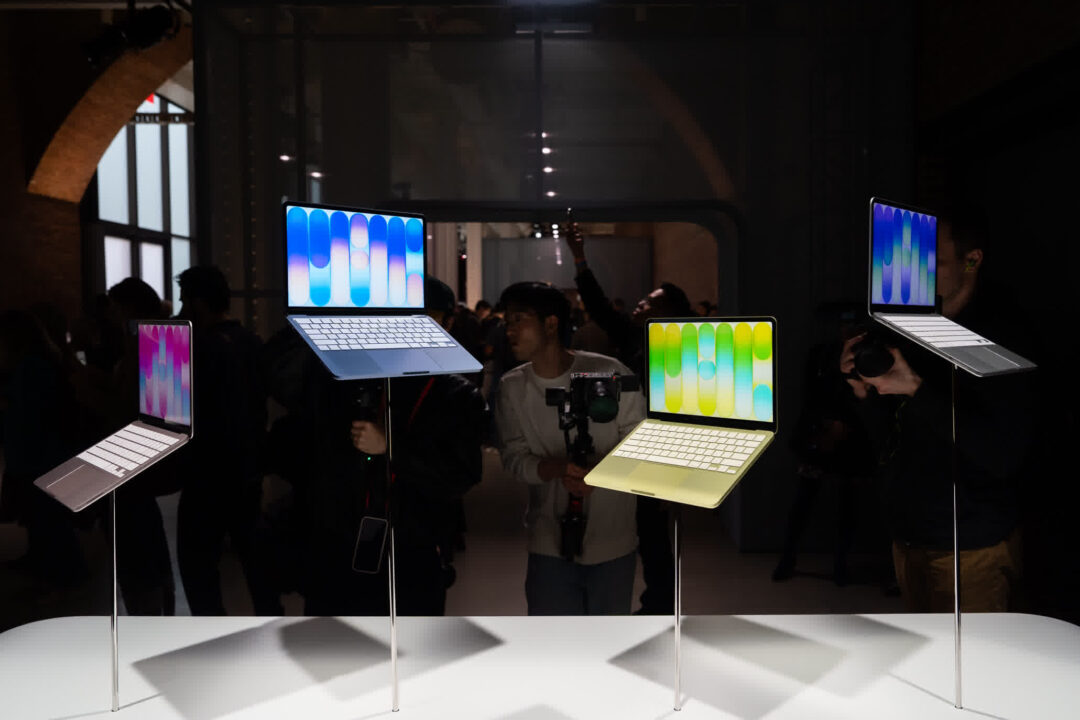 108273477-1772635556615-gettyimages-2264175476-APPLE_LAUNCH_1 Several laptops on tall stands with bright, colorful screens in a dim exhibition space, with visitors in the background.